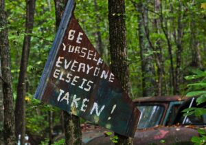 alt= " A poster hanged on trees in the forest written Be yourself every one is taken"